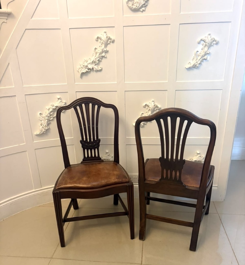 A set of Victorian mahogany Hepplewhite style dining chairs featuring shield-shaped backs with carved splats and tan leather upholstered seats. View X