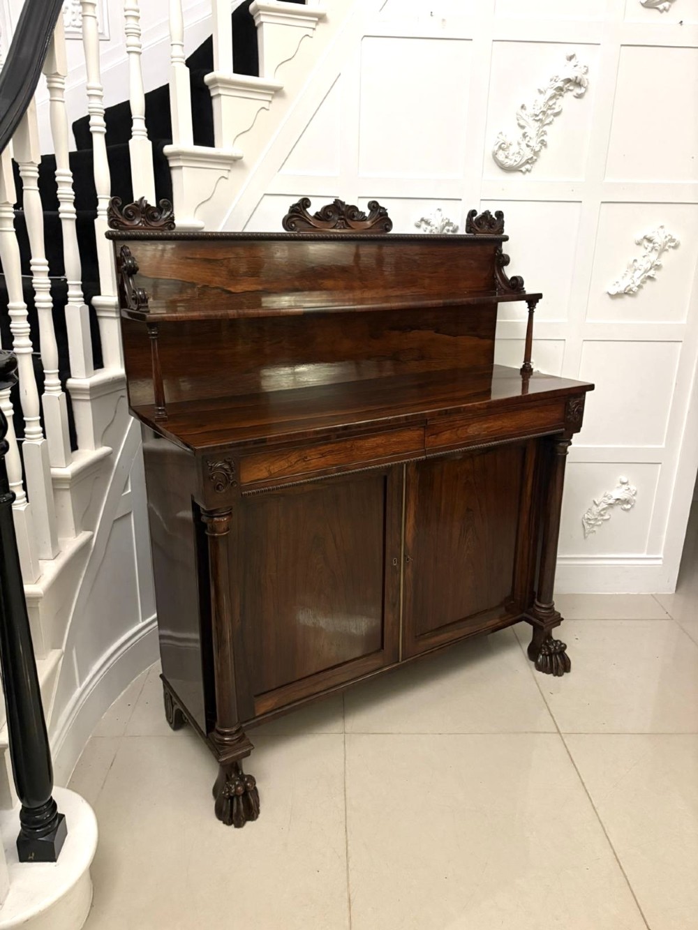 A dark mahogany Regency chiffonier sideboard featuring a two-door lower cupboard, turned support columns, carved claw feet, and a raised upper display shelf with a carved scroll cresting.