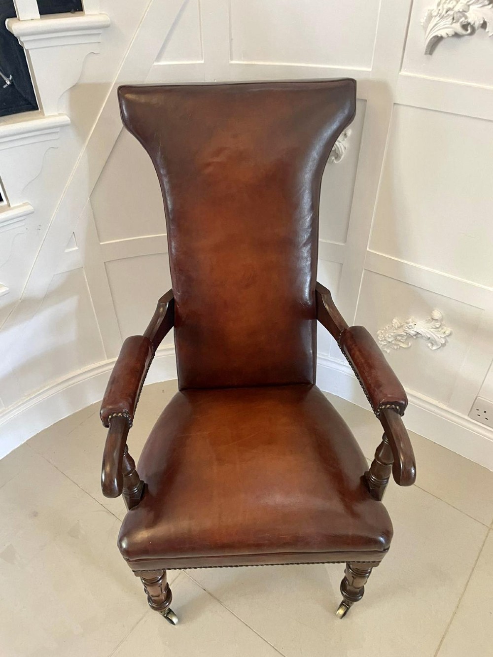 A William IV high-backed armchair upholstered in chestnut brown leather with brass stud detailing, supported by turned mahogany front legs on brass castors. View III