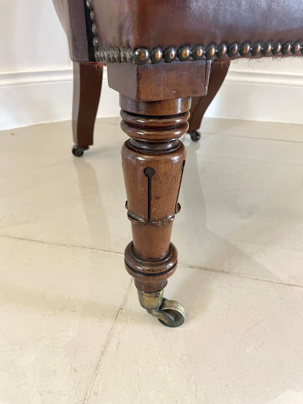 A William IV high-backed armchair upholstered in chestnut brown leather with brass stud detailing, supported by turned mahogany front legs on brass castors. View IV