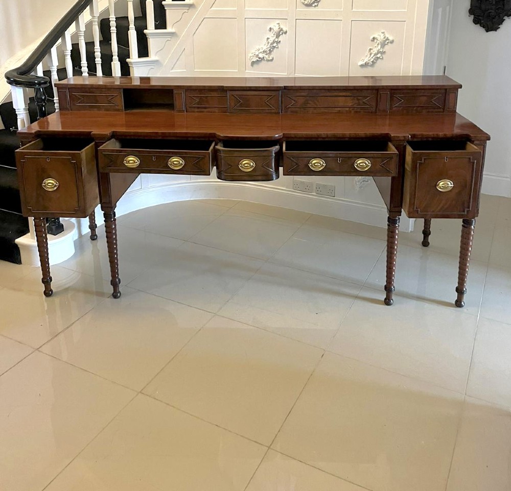 A large George III figured mahogany sideboard featuring a raised gallery, central drawers with brass handles, side cupboards, and turned reeded legs. View VI