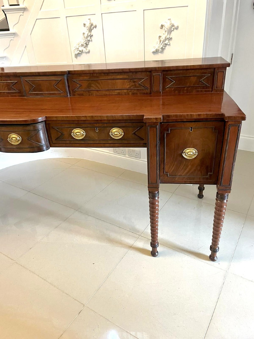 A large George III figured mahogany sideboard featuring a raised gallery, central drawers with brass handles, side cupboards, and turned reeded legs. View XII