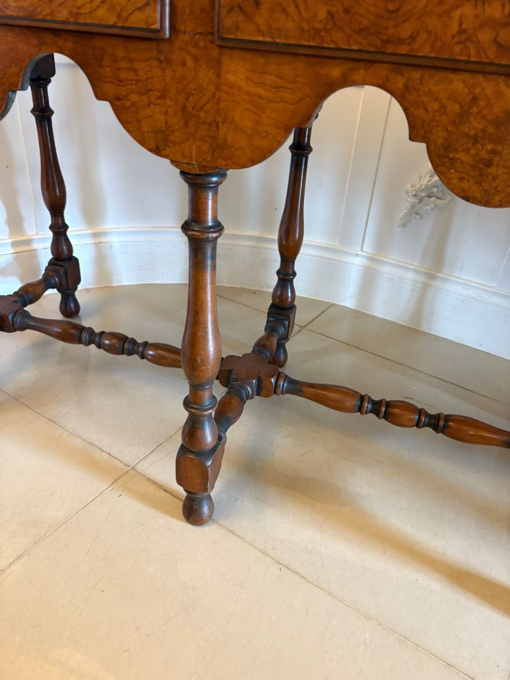 An antique Edwardian burr walnut dresser base featuring three drawers, brass drop handles, a scalloped apron, and turned legs connected by stretchers. View II