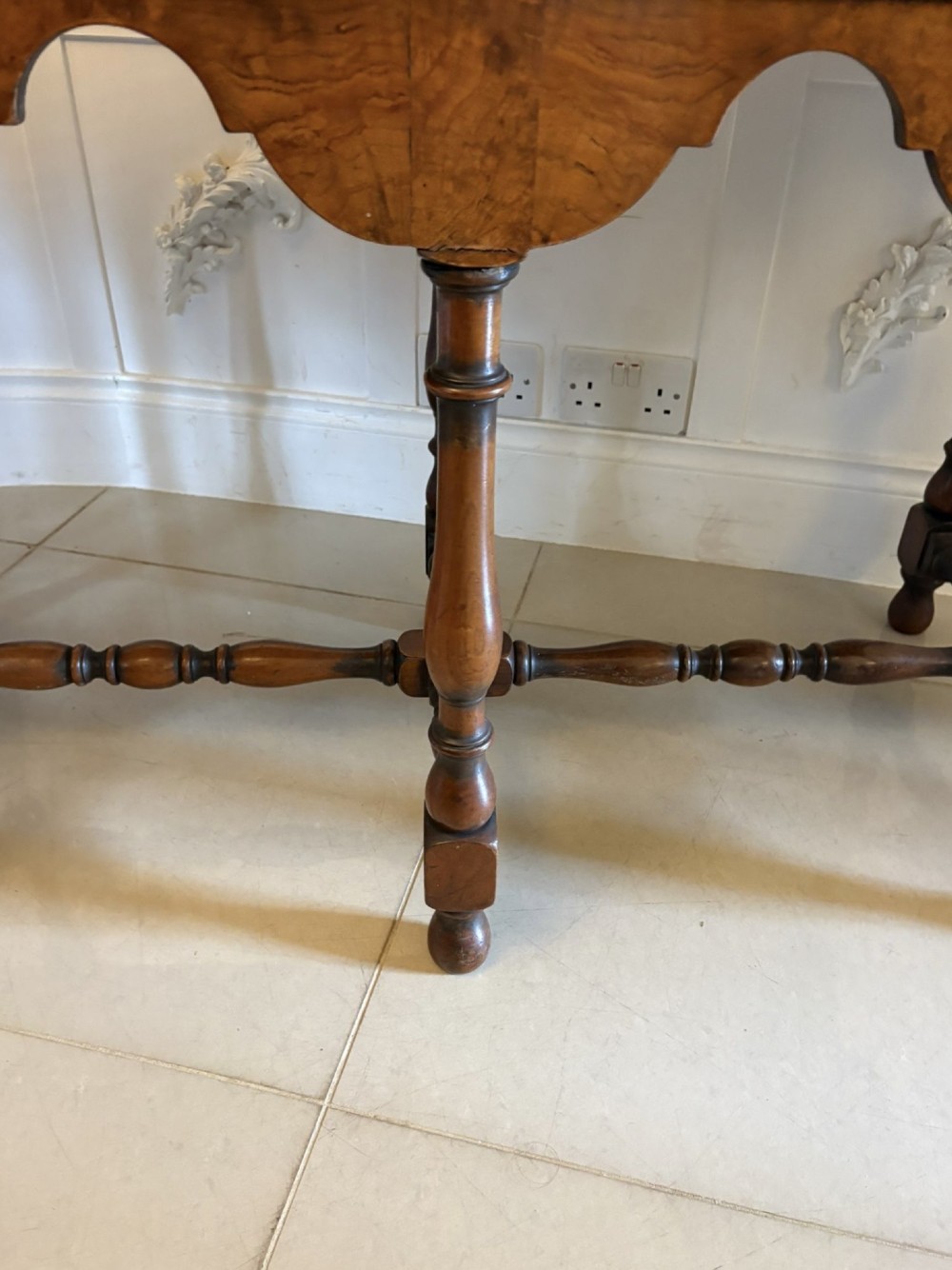 An antique Edwardian burr walnut dresser base featuring three drawers, brass drop handles, a scalloped apron, and turned legs connected by stretchers. View III