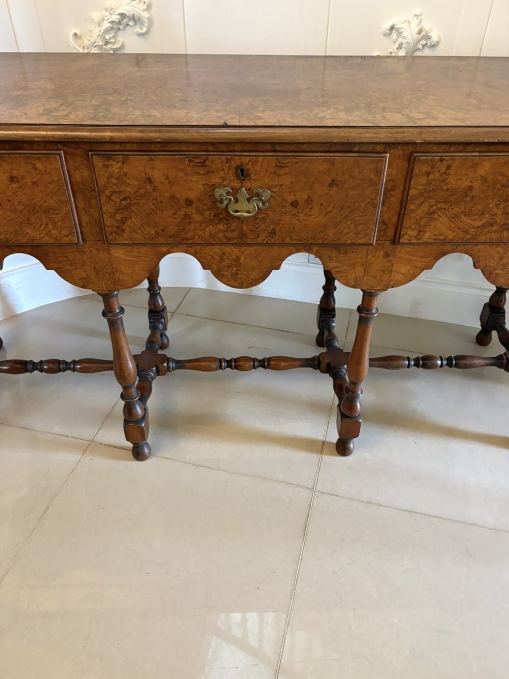 An antique Edwardian burr walnut dresser base featuring three drawers, brass drop handles, a scalloped apron, and turned legs connected by stretchers. View IV
