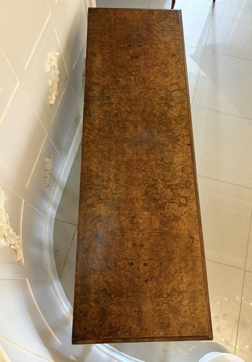 An antique Edwardian burr walnut dresser base featuring three drawers, brass drop handles, a scalloped apron, and turned legs connected by stretchers. View V