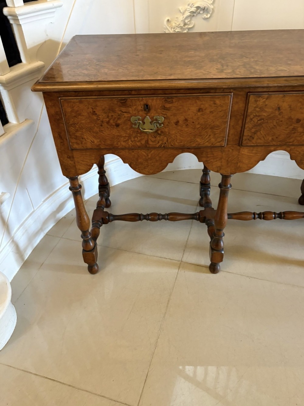 An antique Edwardian burr walnut dresser base featuring three drawers, brass drop handles, a scalloped apron, and turned legs connected by stretchers. View VI