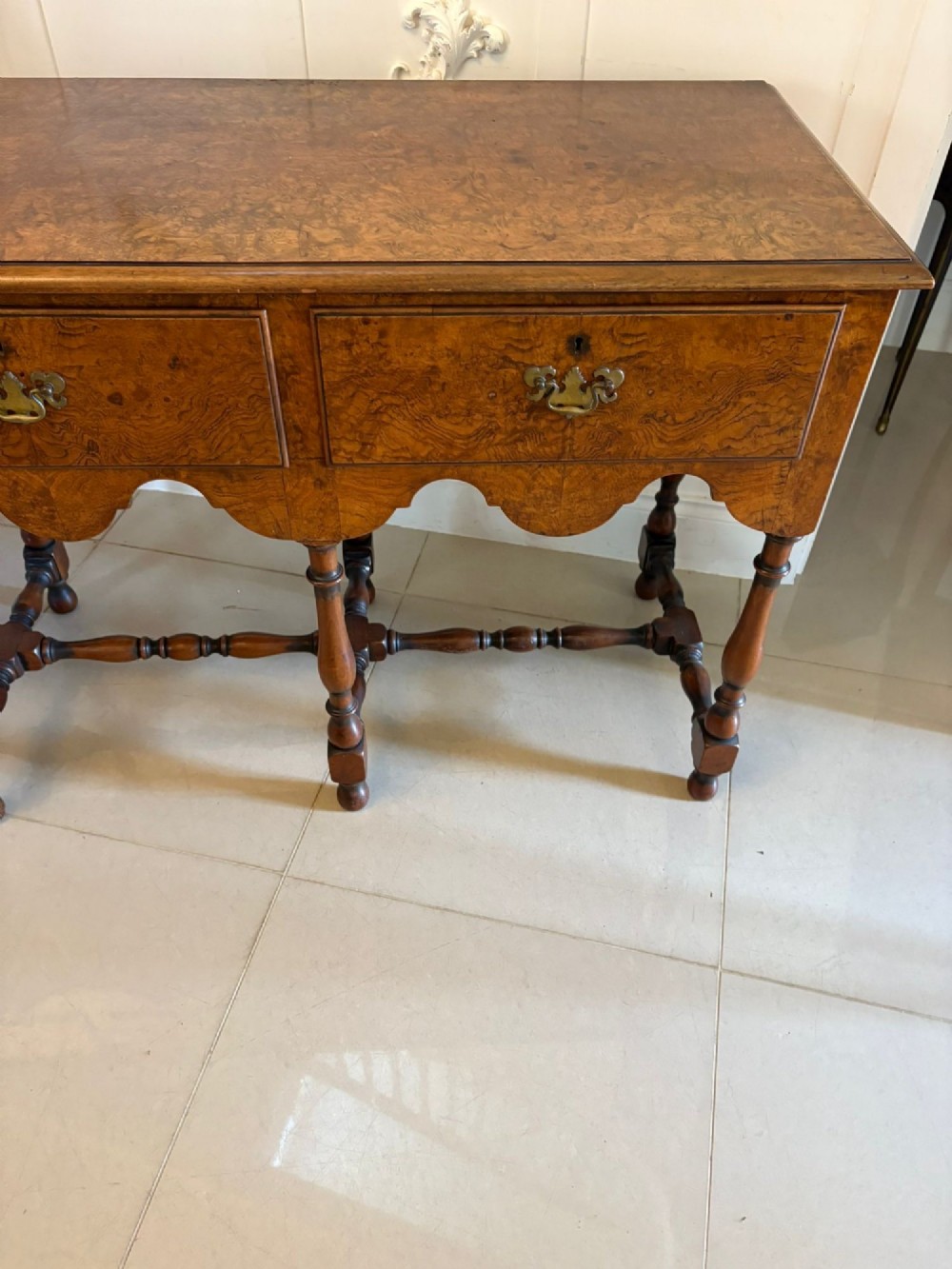 An antique Edwardian burr walnut dresser base featuring three drawers, brass drop handles, a scalloped apron, and turned legs connected by stretchers. View VII