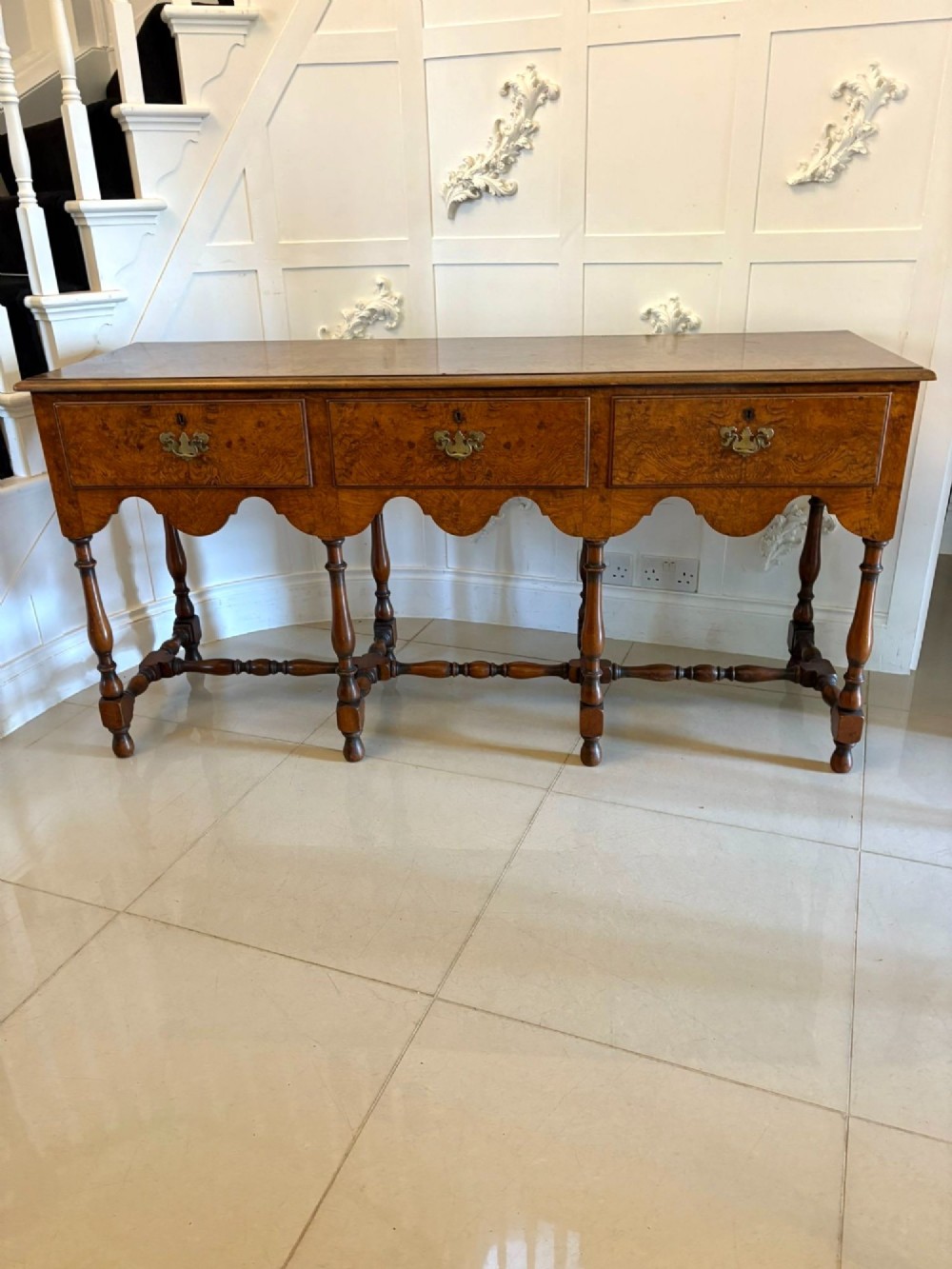 An antique Edwardian burr walnut dresser base featuring three drawers, brass drop handles, a scalloped apron, and turned legs connected by stretchers. View IX