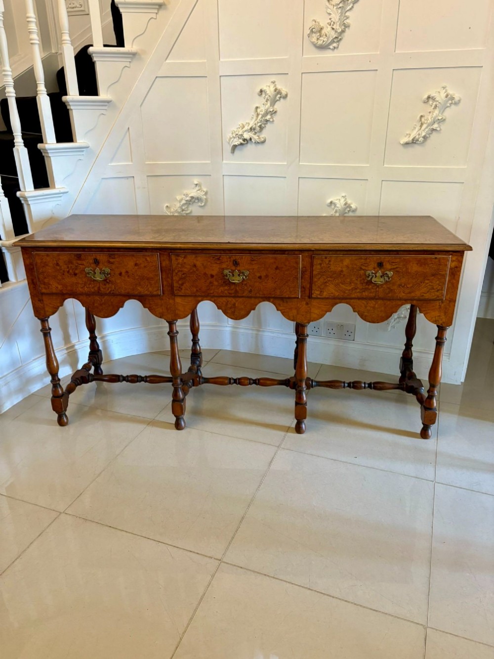 An antique Edwardian burr walnut dresser base featuring three drawers, brass drop handles, a scalloped apron, and turned legs connected by stretchers.