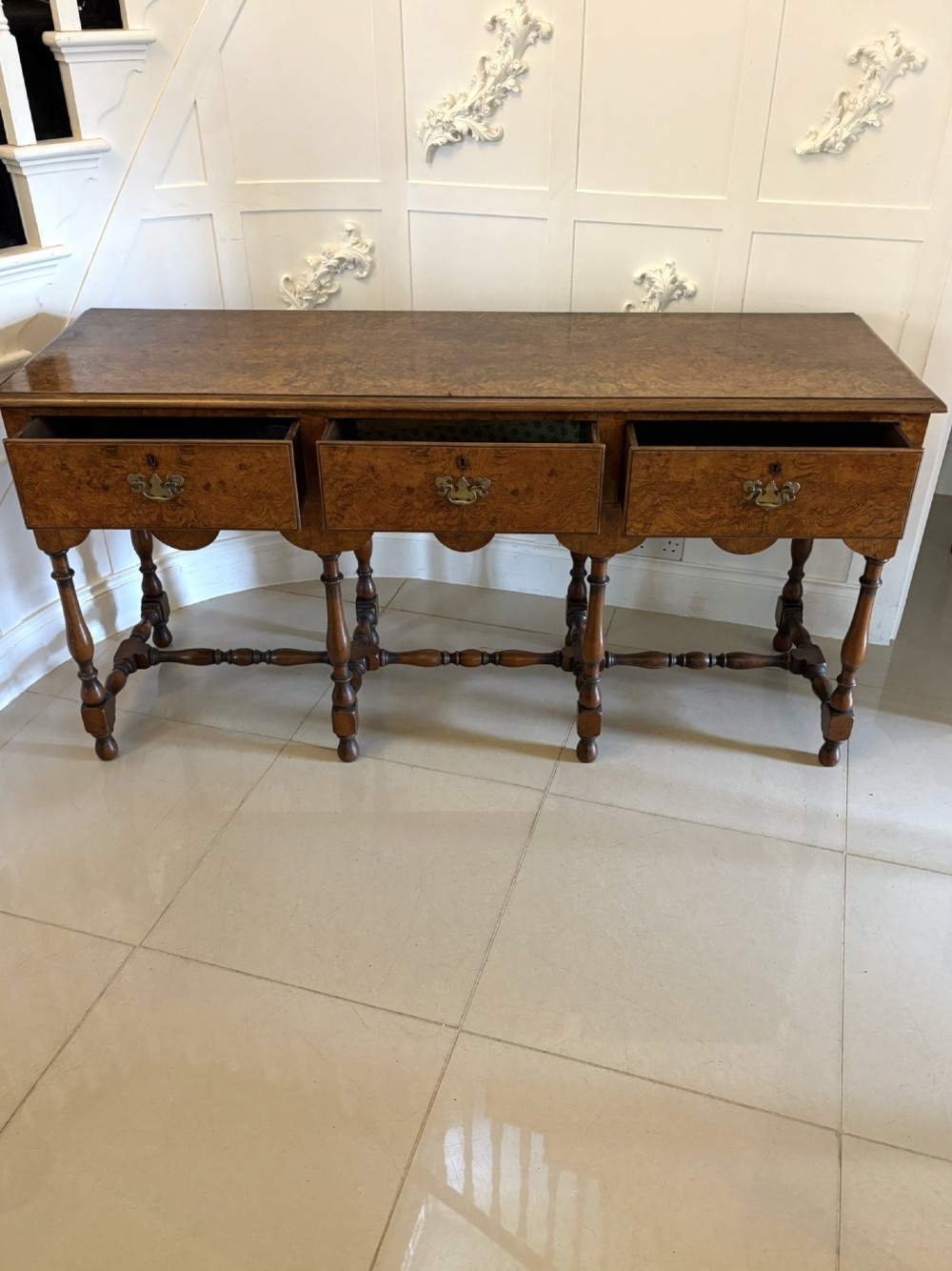 An antique Edwardian burr walnut dresser base featuring three drawers, brass drop handles, a scalloped apron, and turned legs connected by stretchers. View X