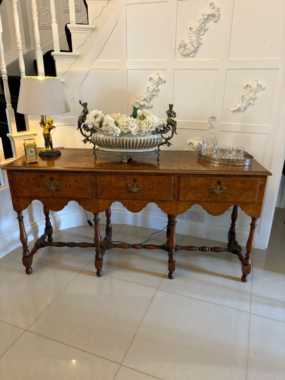 An antique Edwardian burr walnut dresser base featuring three drawers, brass drop handles, a scalloped apron, and turned legs connected by stretchers. View XI