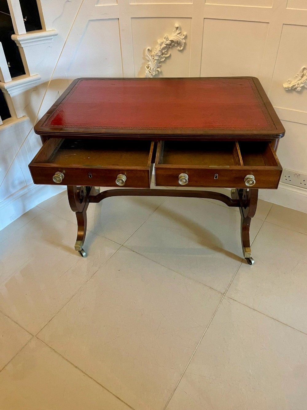 An antique Regency mahogany free-standing writing desk featuring a red leather gilt-tooled insert, two frieze drawers, lyre-form end supports, and brass castors. View IX
