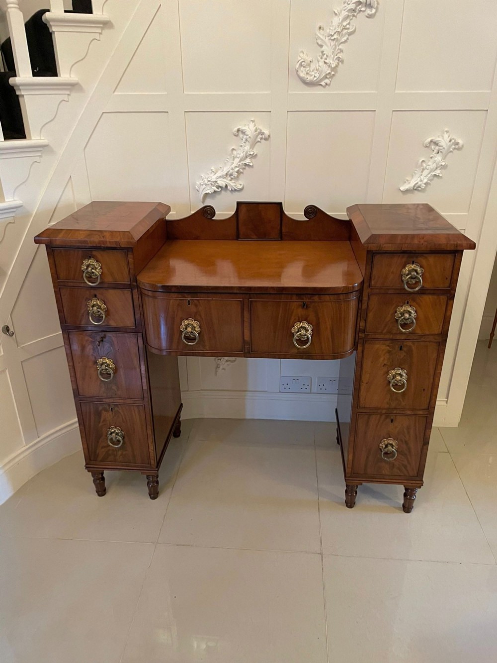 An antique Regency mahogany secretaire sideboard featuring twin pedestals, multiple drawers with brass ring pulls, and a scrolled backboard gallery.