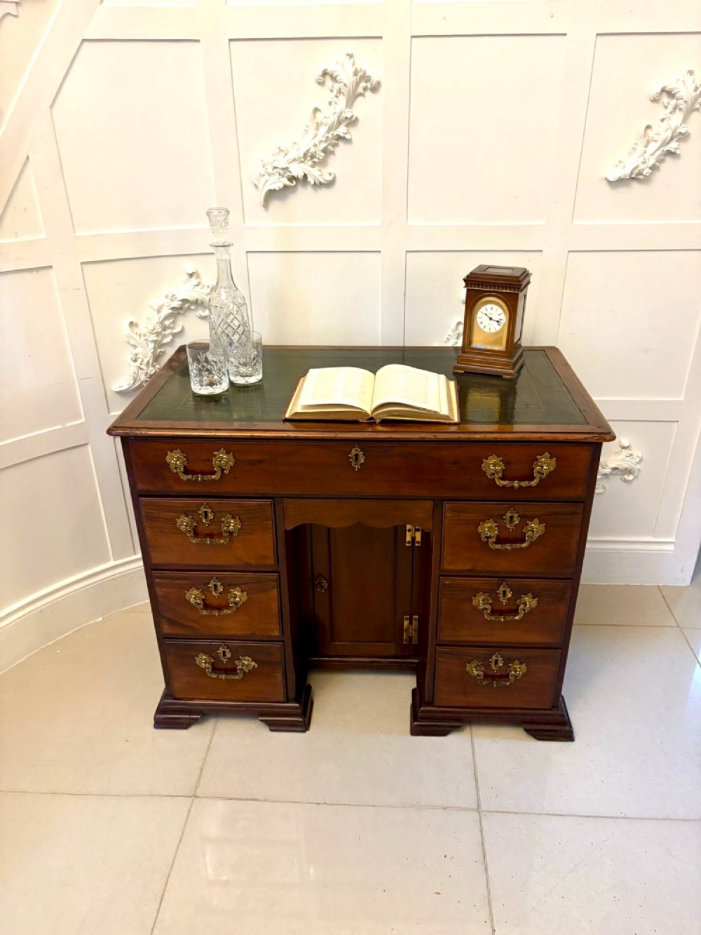 An antique George III mahogany kneehole desk featuring a green leather inset top, brass handles, a central recessed cupboard, and multiple drawers set on bracket feet. View V
