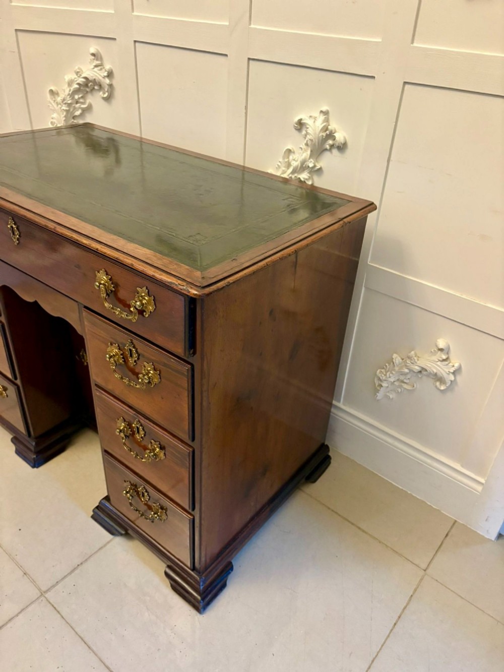 An antique George III mahogany kneehole desk featuring a green leather inset top, brass handles, a central recessed cupboard, and multiple drawers set on bracket feet. View VI