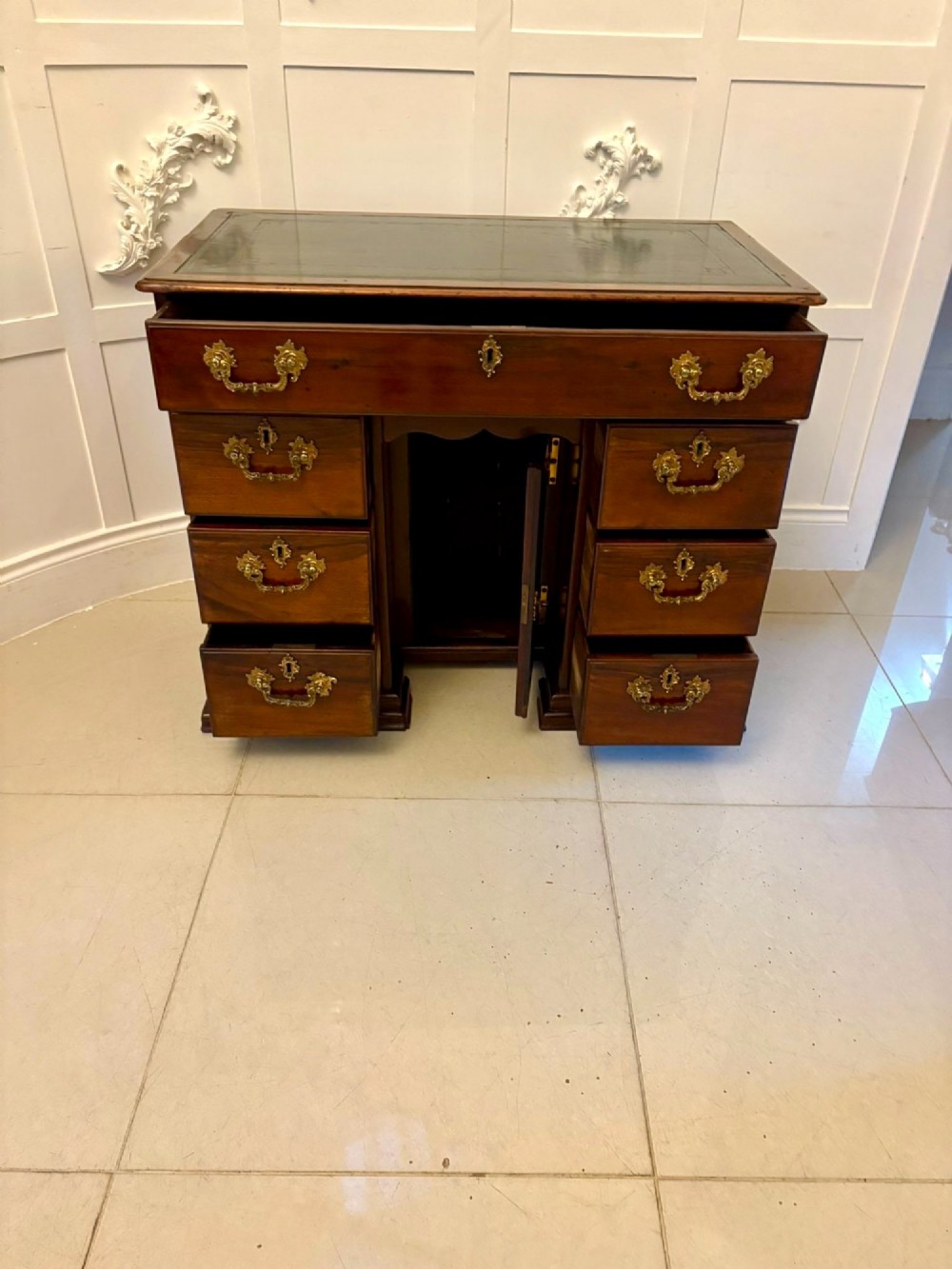 An antique George III mahogany kneehole desk featuring a green leather inset top, brass handles, a central recessed cupboard, and multiple drawers set on bracket feet. View XI