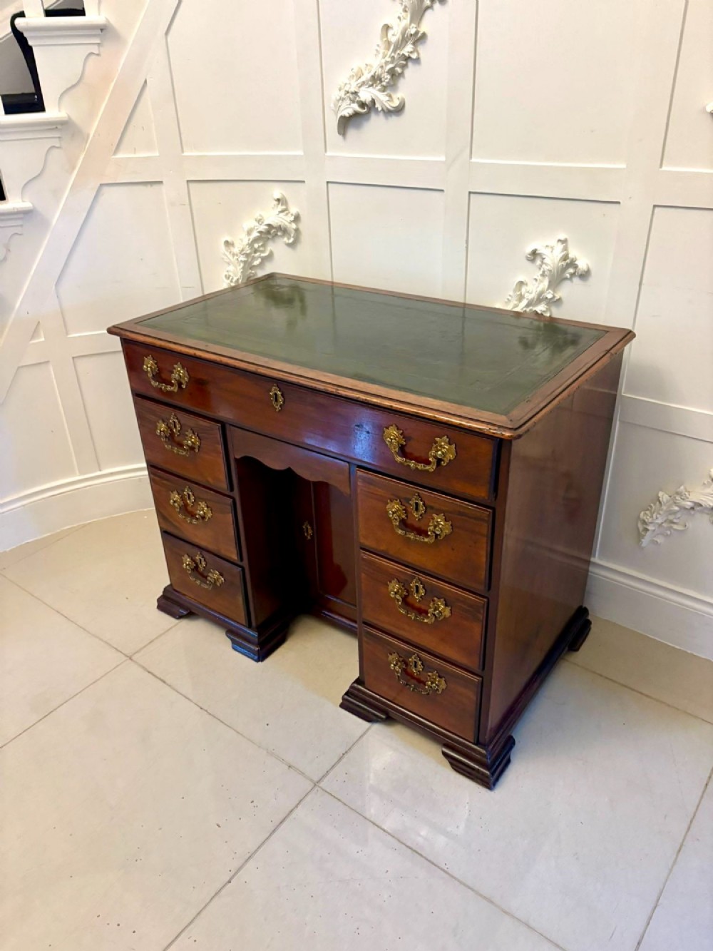 An antique George III mahogany kneehole desk featuring a green leather inset top, brass handles, a central recessed cupboard, and multiple drawers set on bracket feet. View XIII