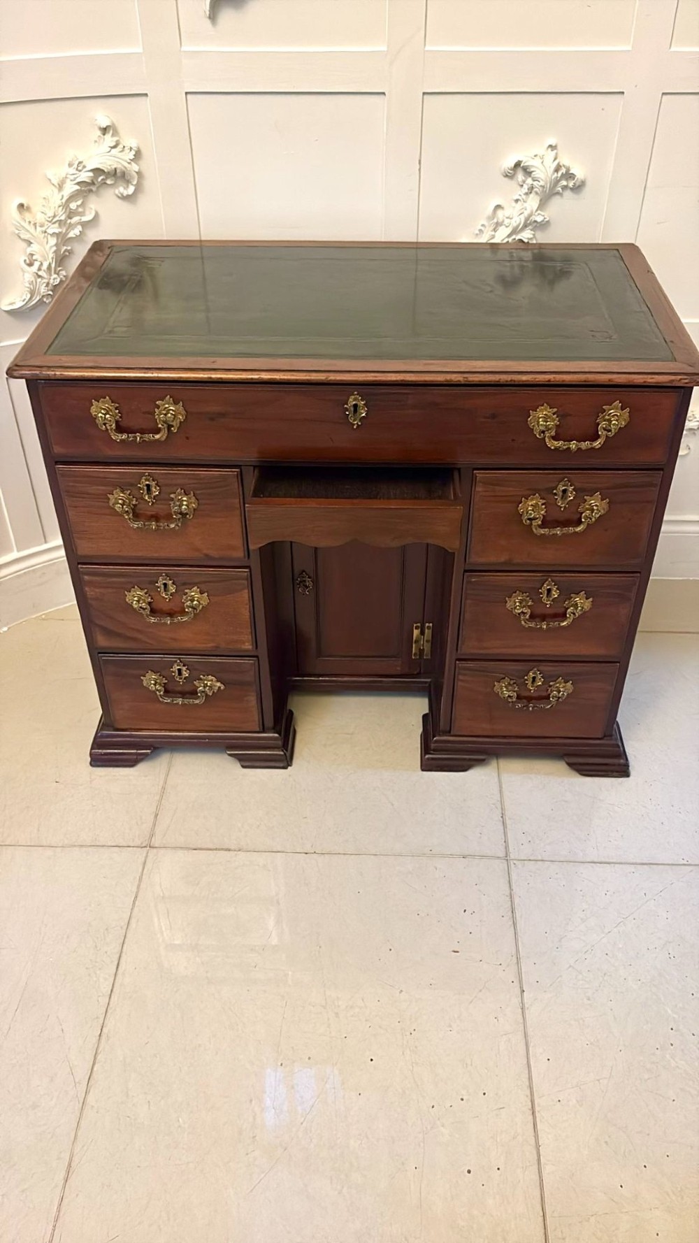 An antique George III mahogany kneehole desk featuring a green leather inset top, brass handles, a central recessed cupboard, and multiple drawers set on bracket feet. View XIV