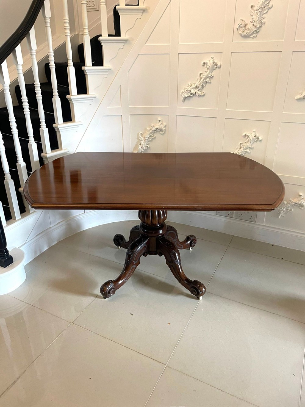 A Victorian mahogany tilt-top dining table with a polished flame mahogany surface shown tilted vertically on a carved pedestal tripod base with castors. View XII
