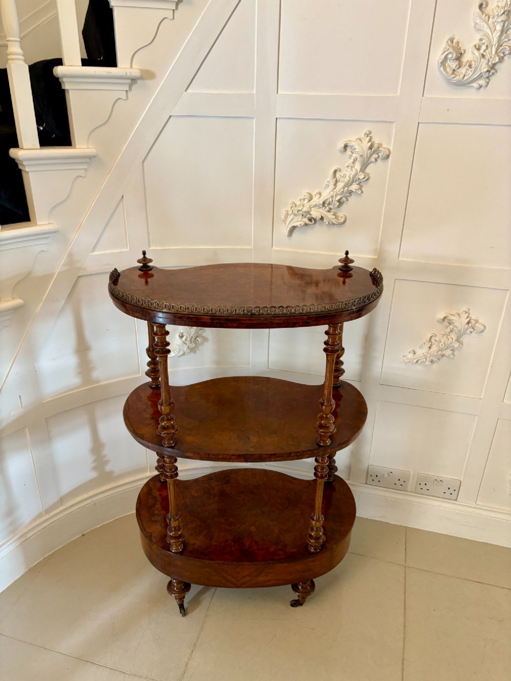 A Victorian burr walnut three-tier serpentine-shaped whatnot featuring turned corner supports, a pierced brass gallery rim, a single bottom drawer, and brass castors. View IV