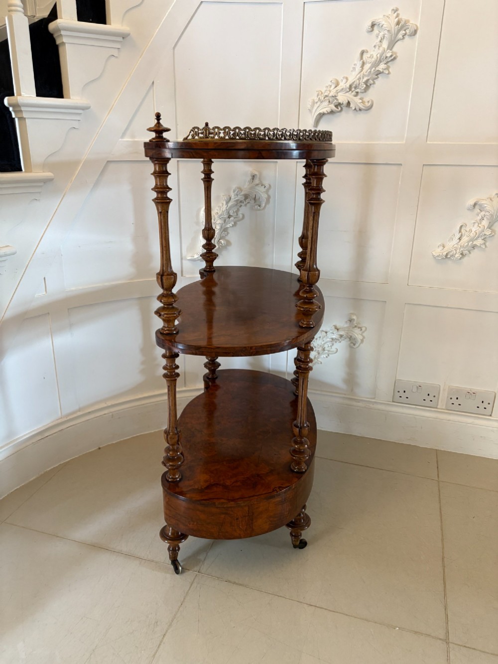 A Victorian burr walnut three-tier serpentine-shaped whatnot featuring turned corner supports, a pierced brass gallery rim, a single bottom drawer, and brass castors. View VI