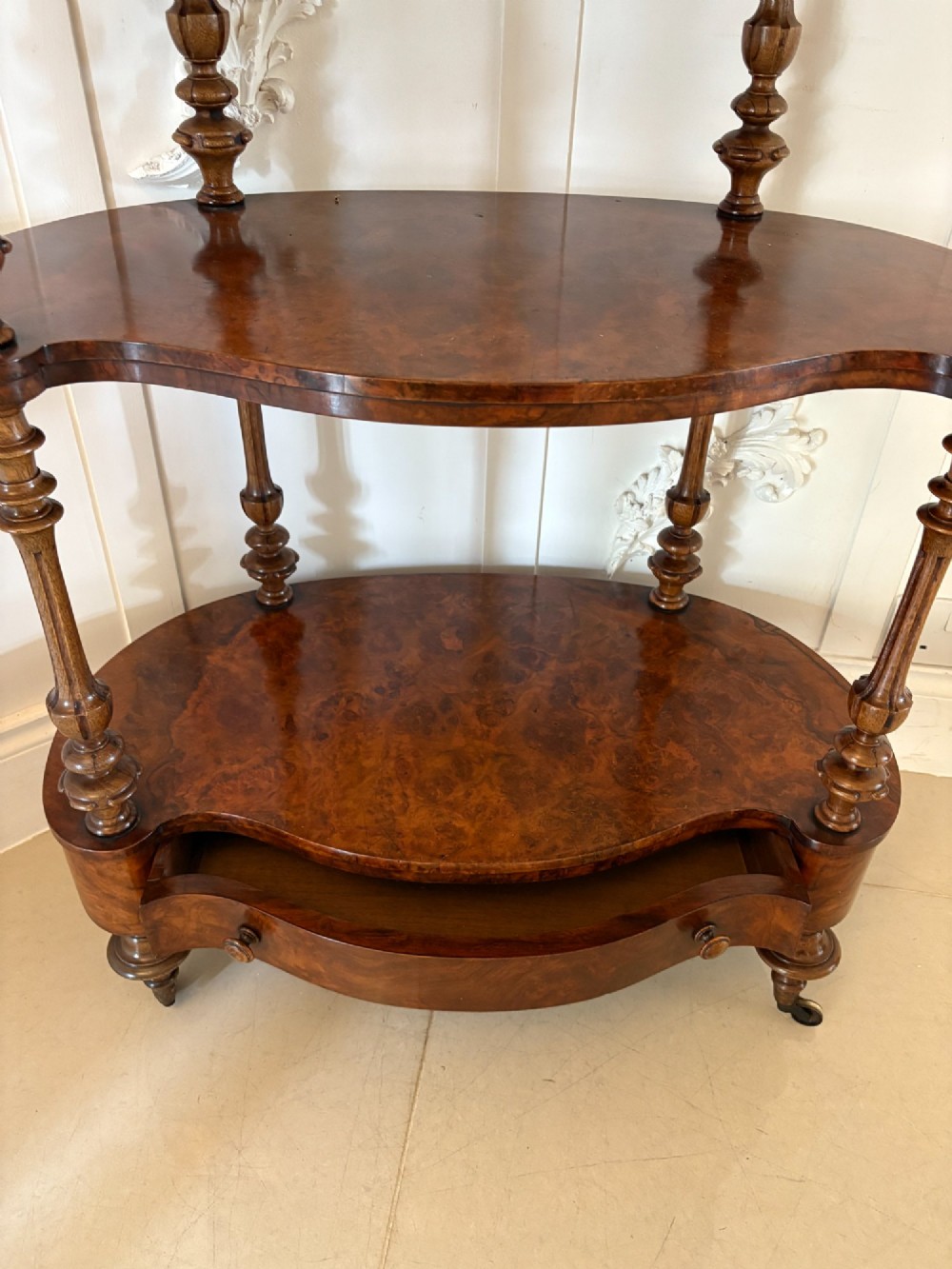 A Victorian burr walnut three-tier serpentine-shaped whatnot featuring turned corner supports, a pierced brass gallery rim, a single bottom drawer, and brass castors. View VII