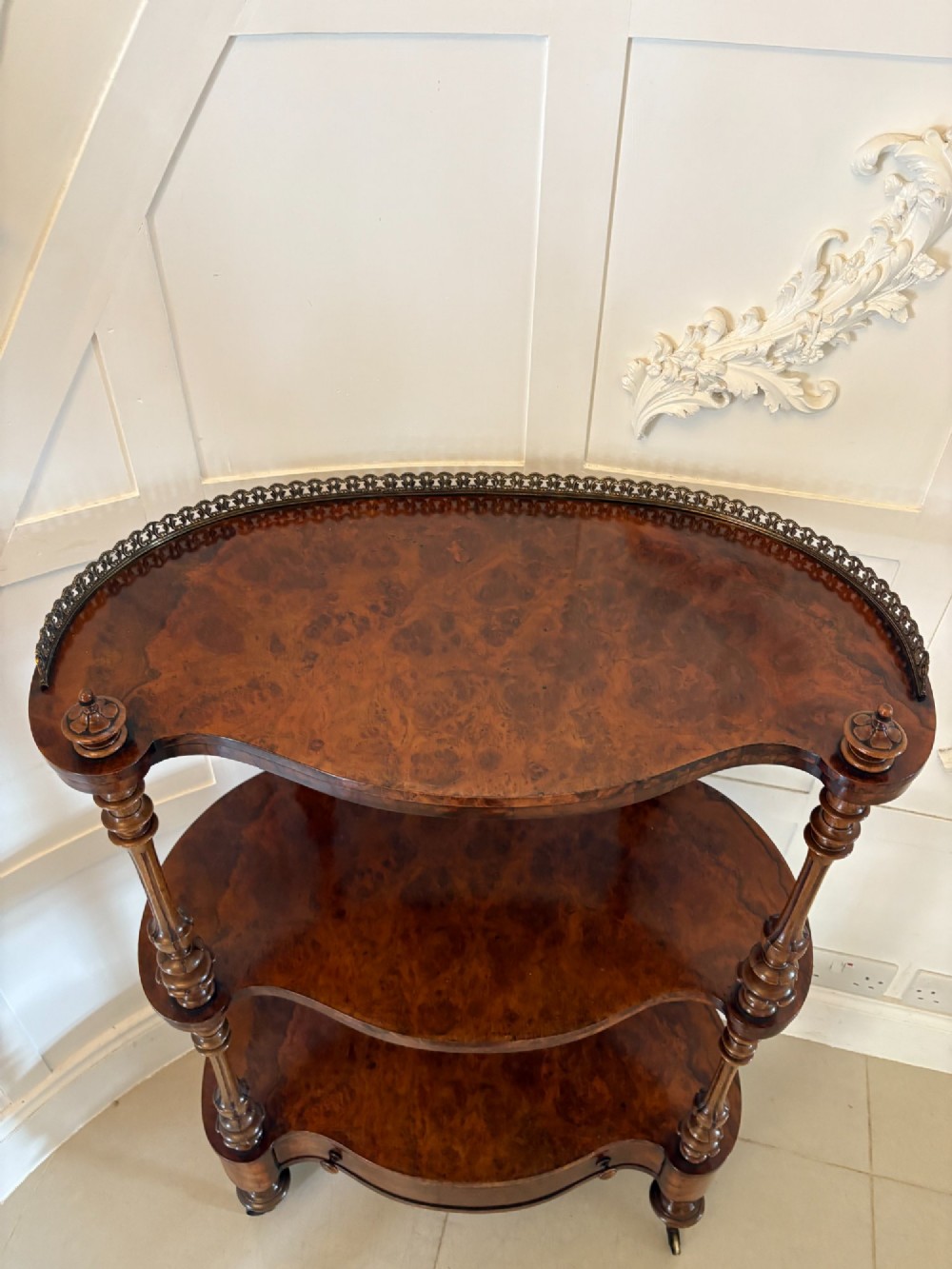 A Victorian burr walnut three-tier serpentine-shaped whatnot featuring turned corner supports, a pierced brass gallery rim, a single bottom drawer, and brass castors. View XIV