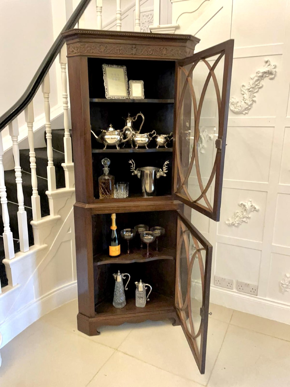 A tall mahogany corner cabinet with two astragal glazed doors featuring curved oval tracery and a carved foliate frieze. View VI