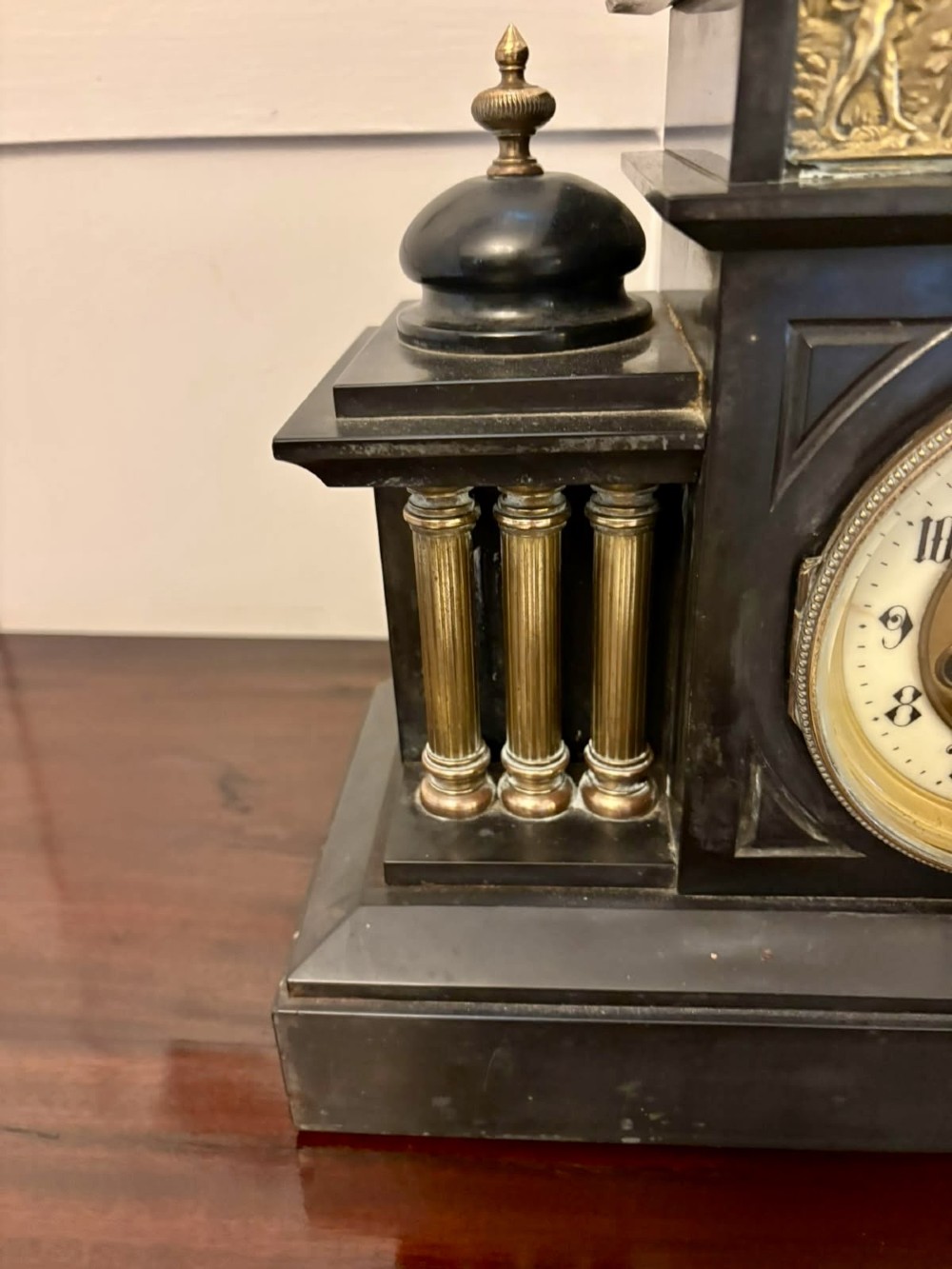 An architectural black marble mantel clock featuring gilt brass columns, a relief frieze panel depicting classical figures, and a round dial with Arabic numerals. View XII