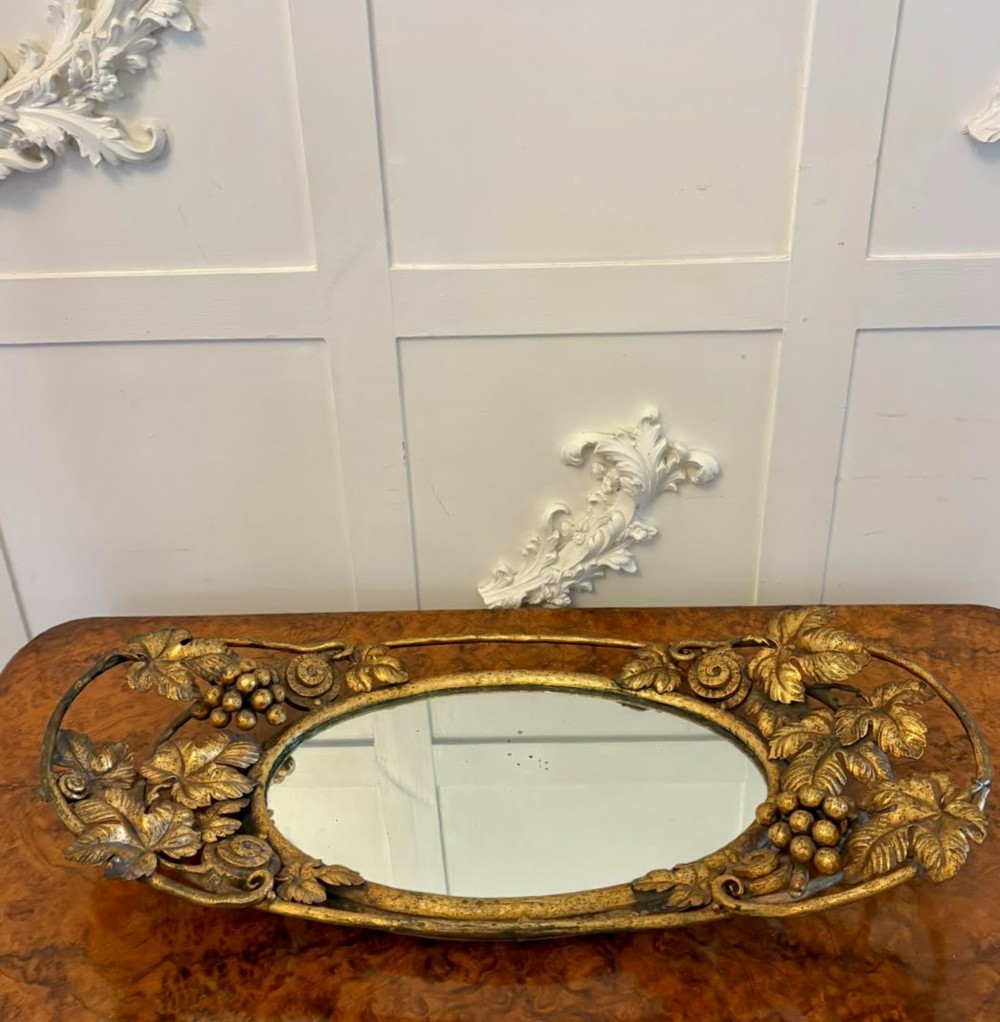 An oval gilt metal tray featuring a central mirror panel surrounded by an ornate border of sculpted grapevines, leaves, and grape clusters.