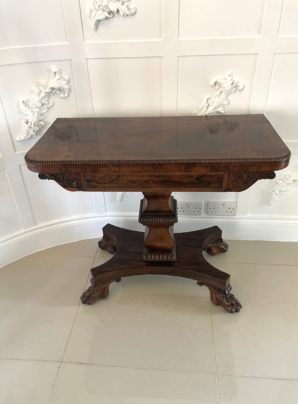 A William IV dark rosewood fold-over card table featuring a gadrooned top edge, scroll carvings, a central pedestal column, and a plinth base on carved claw feet. View II