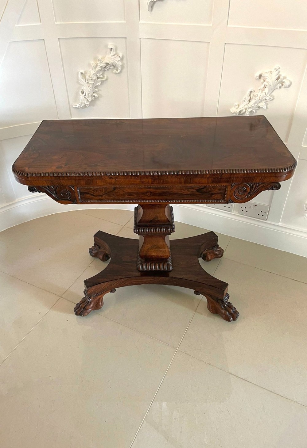 A William IV dark rosewood fold-over card table featuring a gadrooned top edge, scroll carvings, a central pedestal column, and a plinth base on carved claw feet. View III
