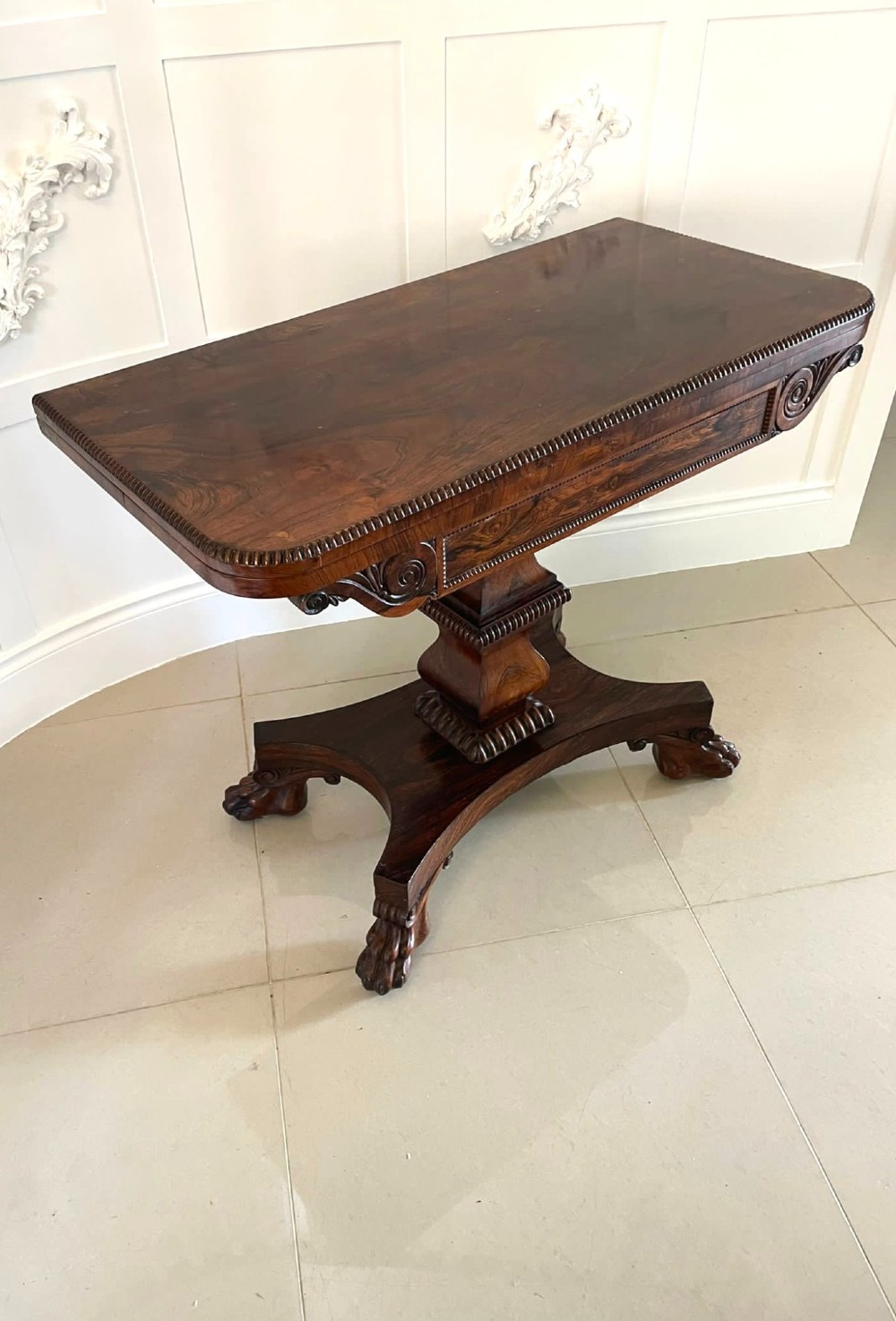 A William IV dark rosewood fold-over card table featuring a gadrooned top edge, scroll carvings, a central pedestal column, and a plinth base on carved claw feet.