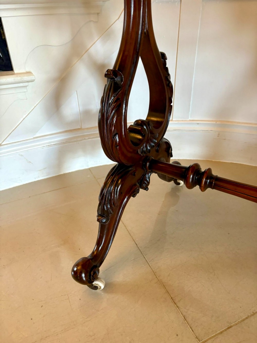 A Victorian burr walnut centre table with a serpentine shaped top, figured wood grain, detailed carved end supports, and a turned stretcher on small castors. View II