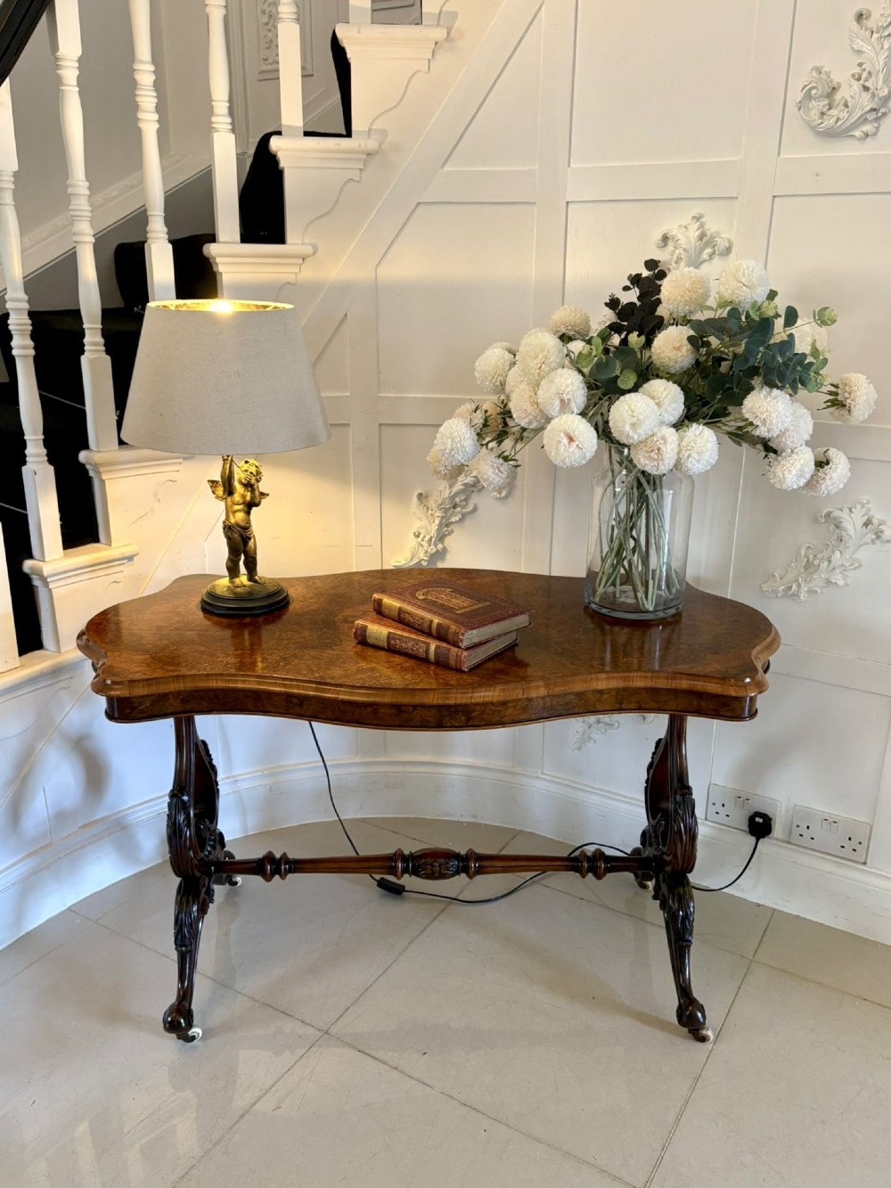 A Victorian burr walnut centre table with a serpentine shaped top, figured wood grain, detailed carved end supports, and a turned stretcher on small castors. View IV
