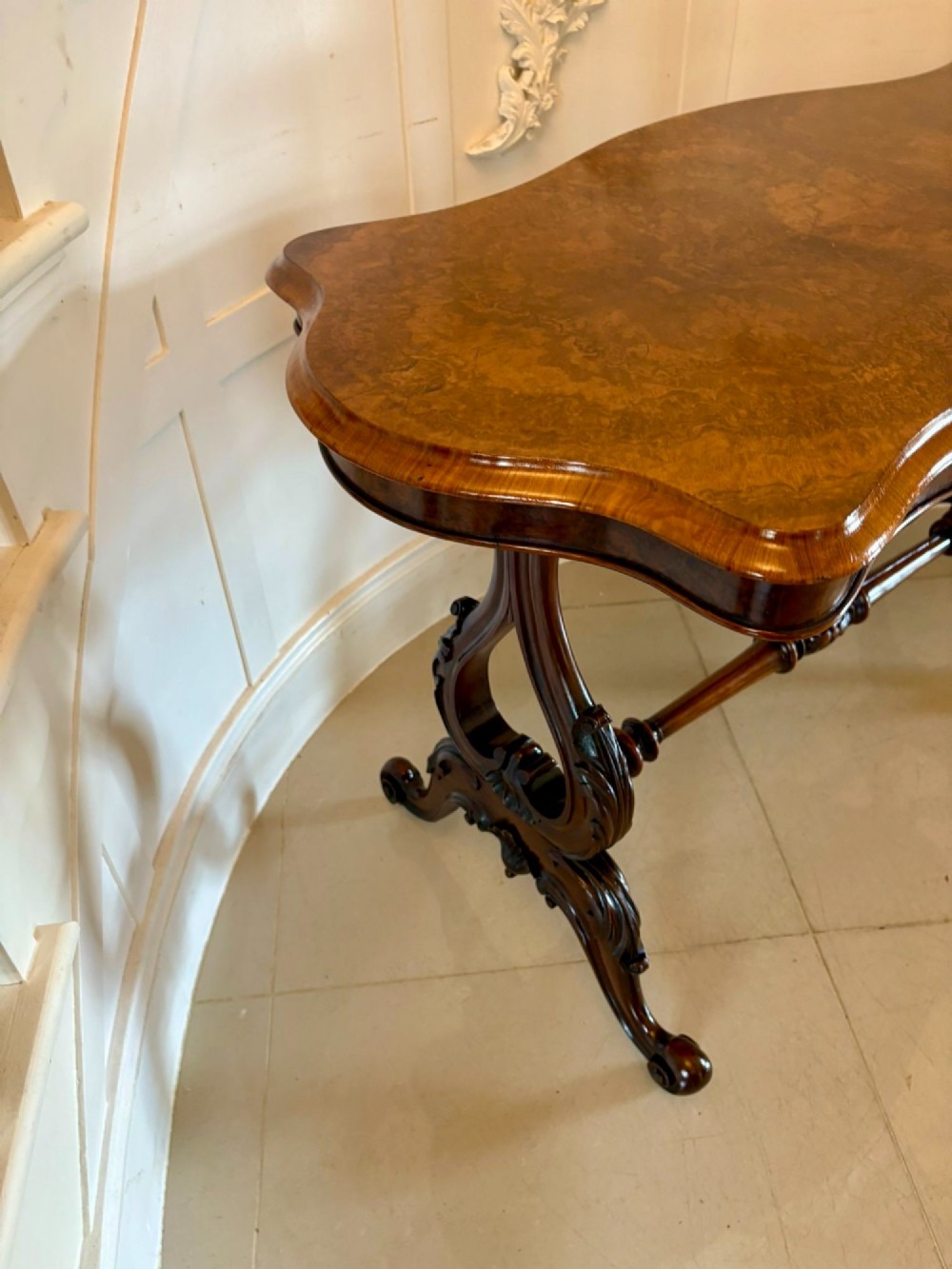 A Victorian burr walnut centre table with a serpentine shaped top, figured wood grain, detailed carved end supports, and a turned stretcher on small castors. View V