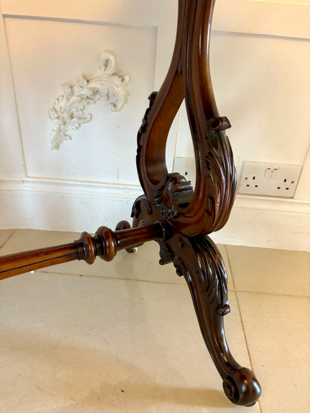 A Victorian burr walnut centre table with a serpentine shaped top, figured wood grain, detailed carved end supports, and a turned stretcher on small castors. View VII