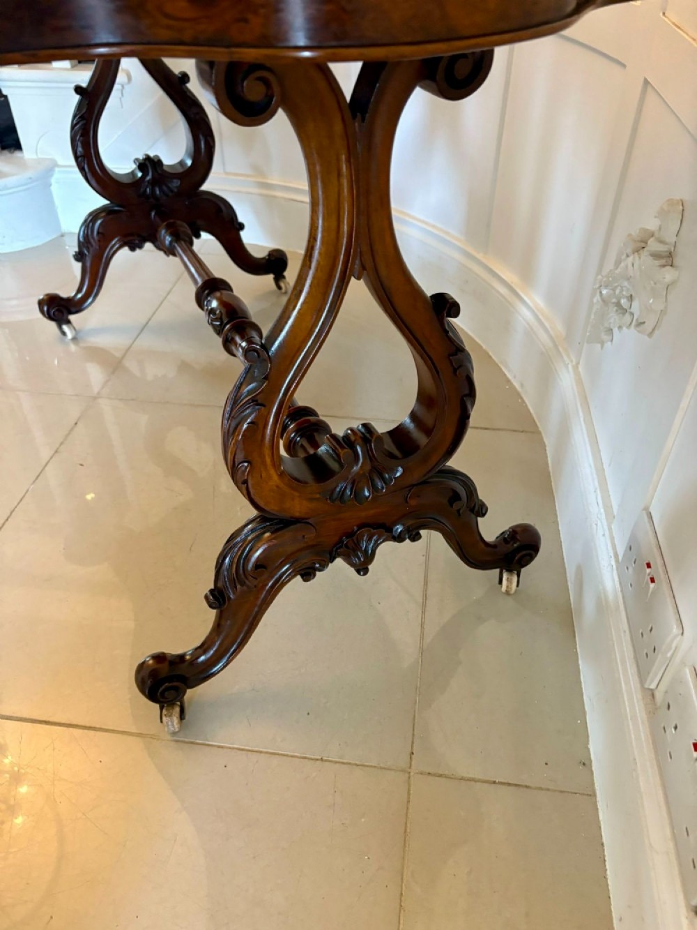 A Victorian burr walnut centre table with a serpentine shaped top, figured wood grain, detailed carved end supports, and a turned stretcher on small castors. View VIII