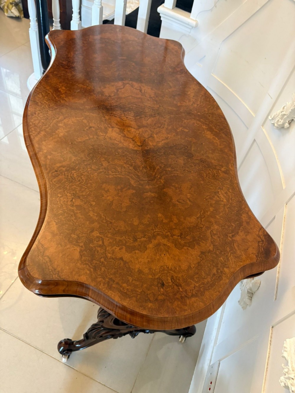 A Victorian burr walnut centre table with a serpentine shaped top, figured wood grain, detailed carved end supports, and a turned stretcher on small castors. View IX