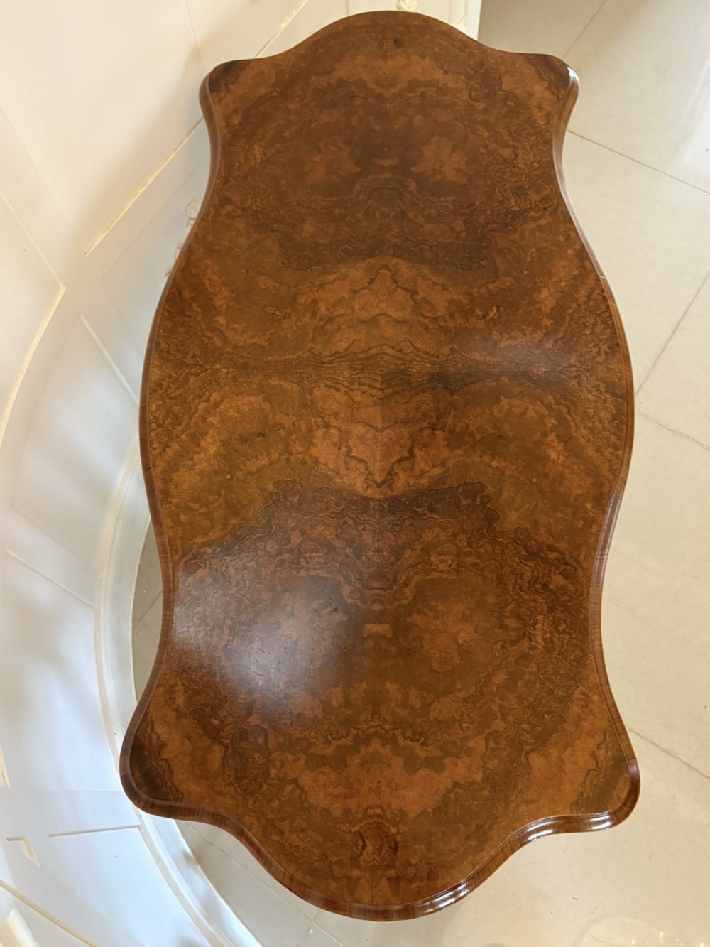 A Victorian burr walnut centre table with a serpentine shaped top, figured wood grain, detailed carved end supports, and a turned stretcher on small castors. View X