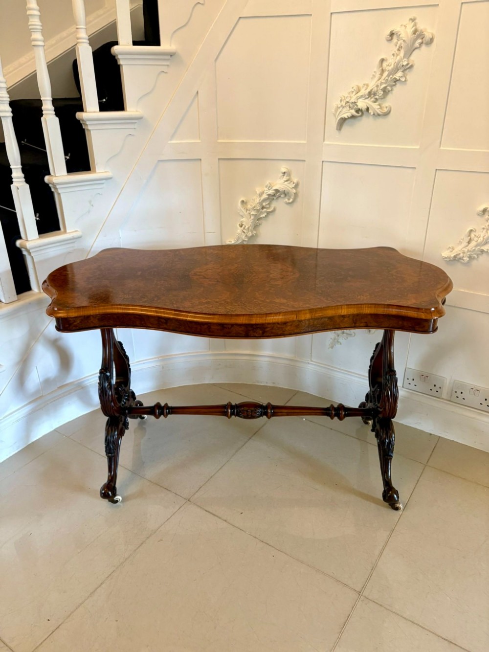 A Victorian burr walnut centre table with a serpentine shaped top, figured wood grain, detailed carved end supports, and a turned stretcher on small castors.