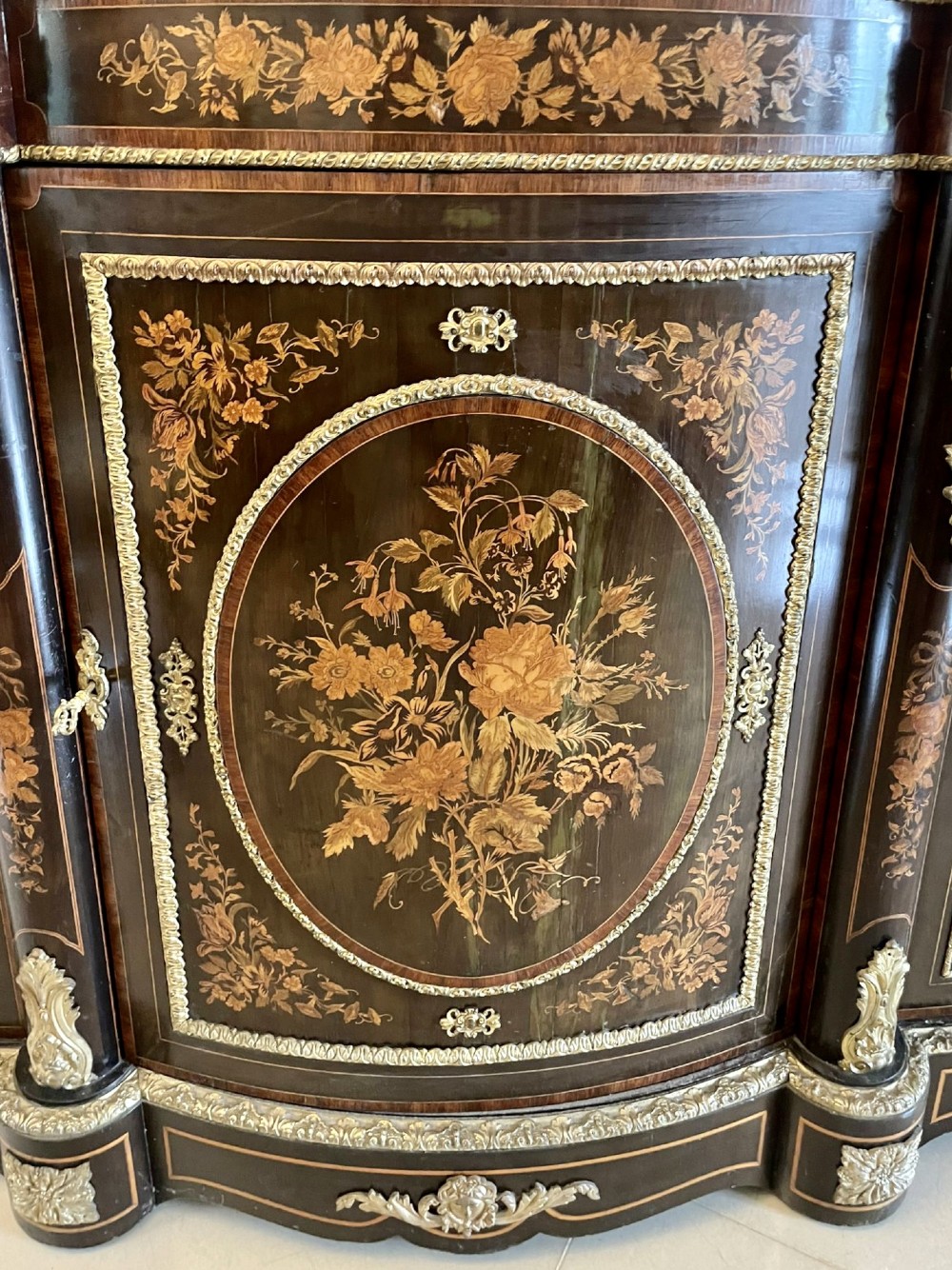 A dark wood Victorian serpentine credenza with floral marquetry inlay, gilt ormolu mounts, and two curved glass display doors. View VII