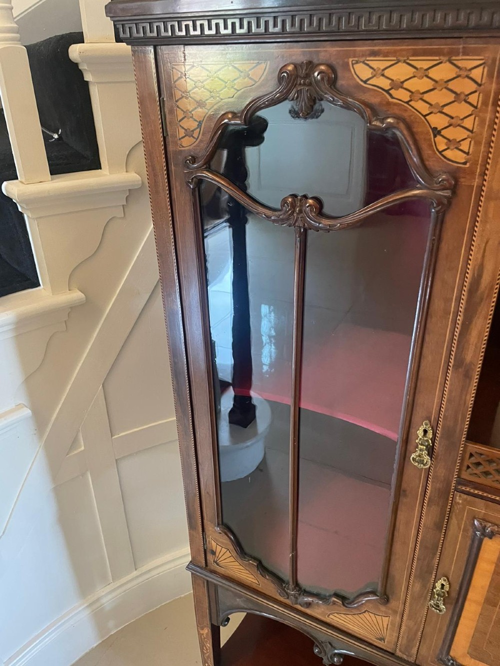 A large Victorian mahogany display cabinet featuring a carved pediment top, central bevelled mirror back, floral satinwood marquetry panel, glazed doors, and a bottom shelf. View II