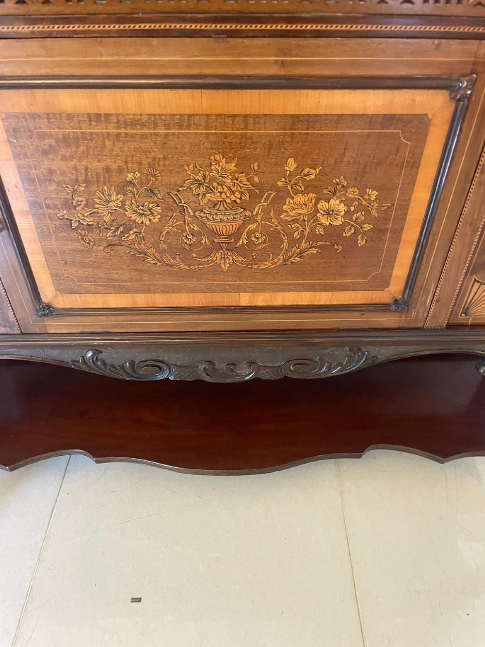 A large Victorian mahogany display cabinet featuring a carved pediment top, central bevelled mirror back, floral satinwood marquetry panel, glazed doors, and a bottom shelf. View V