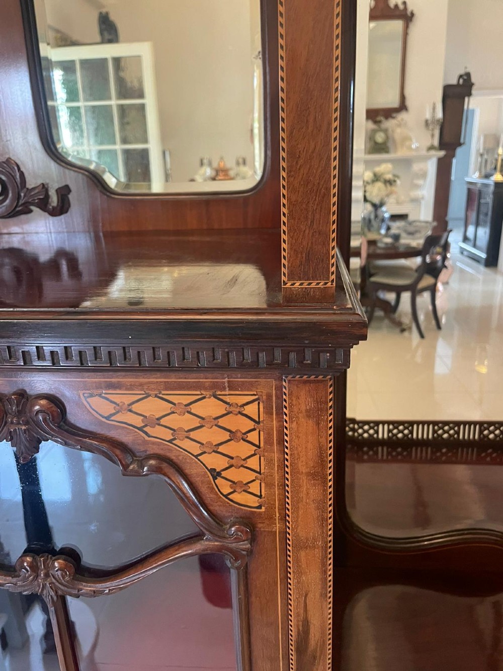 A large Victorian mahogany display cabinet featuring a carved pediment top, central bevelled mirror back, floral satinwood marquetry panel, glazed doors, and a bottom shelf. View VI