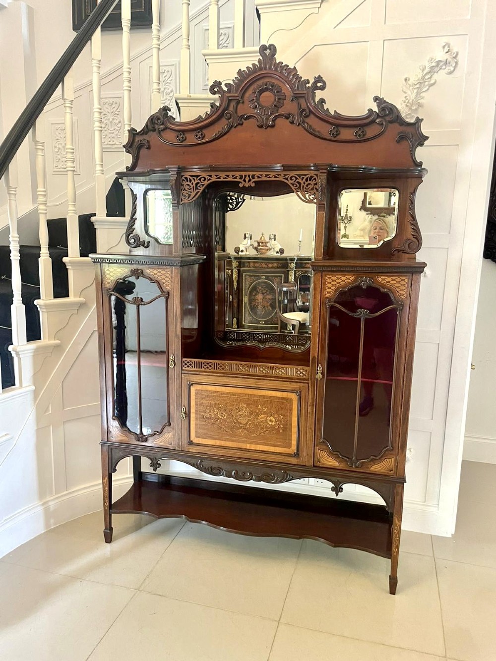 A large Victorian mahogany display cabinet featuring a carved pediment top, central bevelled mirror back, floral satinwood marquetry panel, glazed doors, and a bottom shelf. View XI