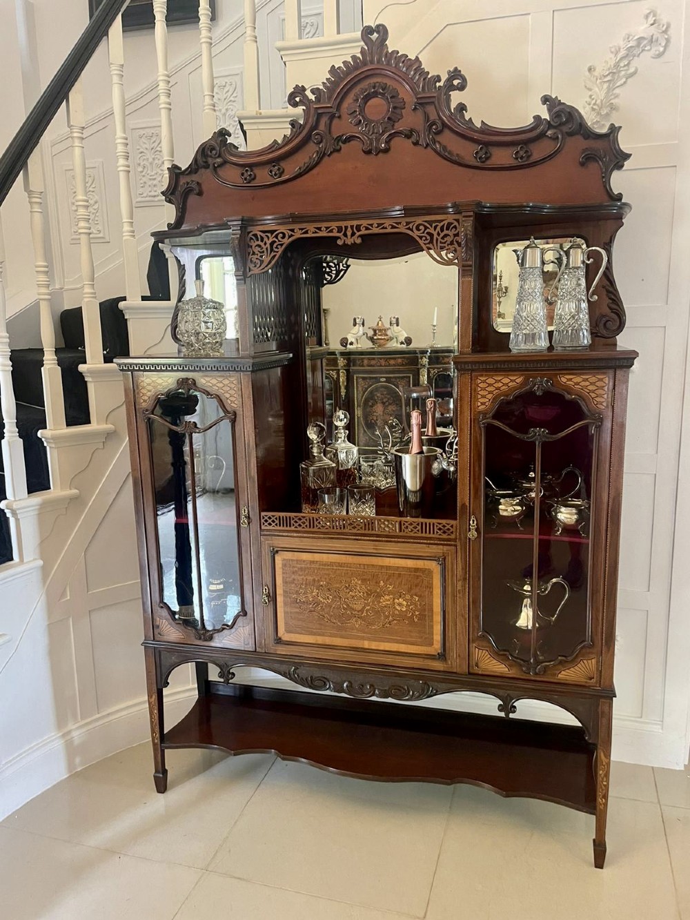 A large Victorian mahogany display cabinet featuring a carved pediment top, central bevelled mirror back, floral satinwood marquetry panel, glazed doors, and a bottom shelf. View XIII