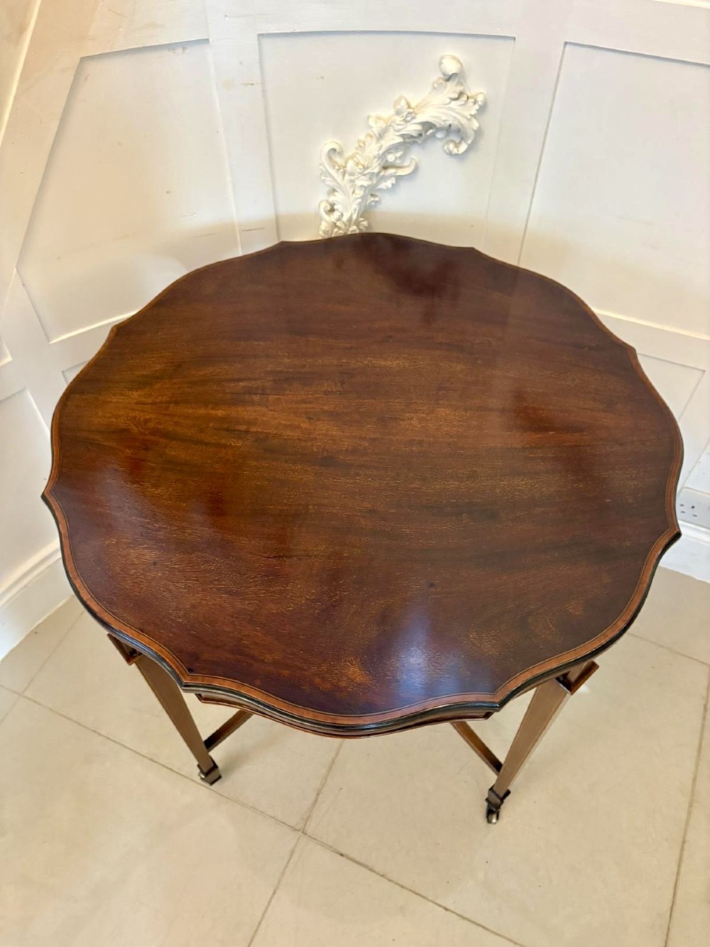 An antique Edwardian polished wood centre table featuring a shaped scalloped top, satinwood inlaid banding, tapered legs with brass castors, and an X-frame stretcher with a central finial. View III