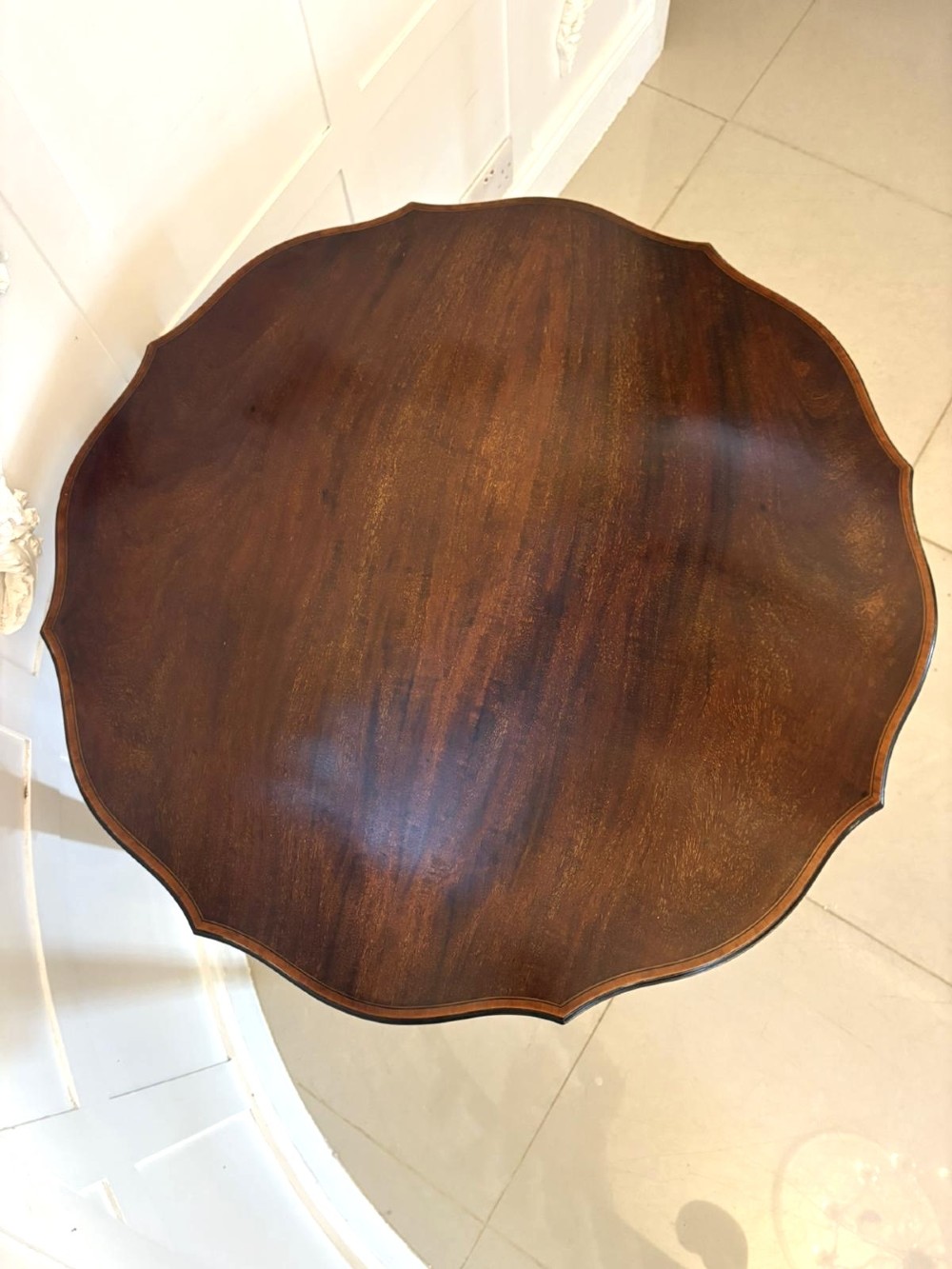 An antique Edwardian polished wood centre table featuring a shaped scalloped top, satinwood inlaid banding, tapered legs with brass castors, and an X-frame stretcher with a central finial. View IV